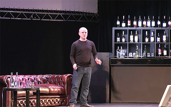 Marco Arment presents a conference talk at NSConference, on a stage designed like a whisky bar with an ornate leather couch.
